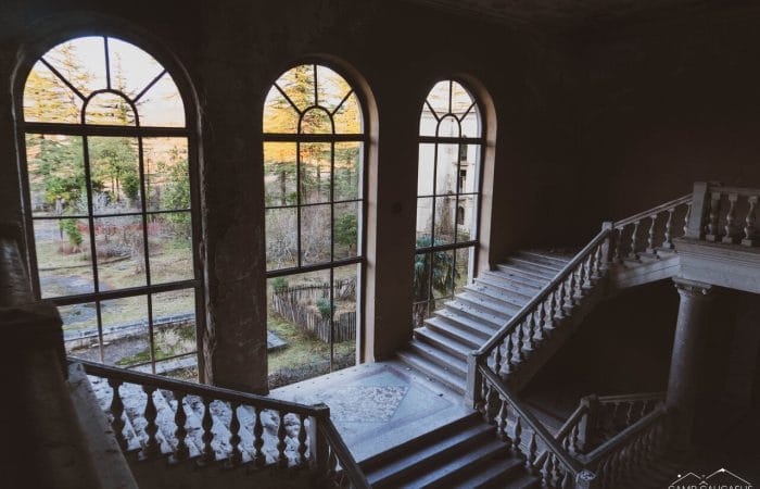 Interior view of an abandoned Soviet-era sanatorium in Tskaltubo, Georgia, featuring a grand staircase and large arched windows with sunlight illuminating the decaying architecture