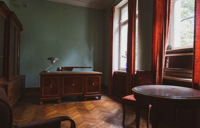 Vintage wooden desk, table, and red velvet chairs inside an abandoned Soviet-era sanatorium room with green walls and sunlight streaming through tall windows in Tskaltubo, Georgia — urbex photography, historic interior, post-Soviet architecture