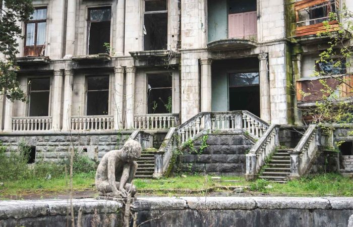 Weathered stone statue sitting beside an empty fountain in front of an abandoned Soviet-era sanatorium with broken windows and crumbling facade in Tskaltubo, Georgia — historic architecture, urbex travel, and post-Soviet heritage