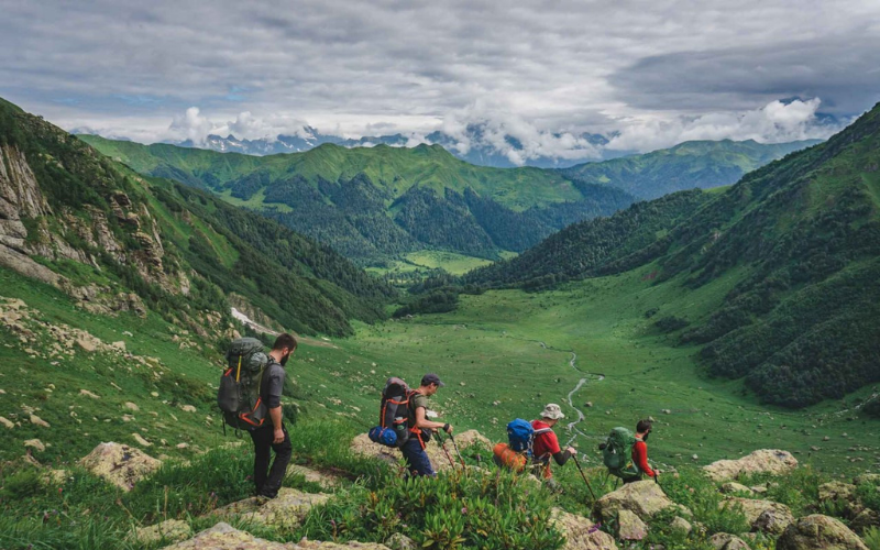 A group of hikers with backpacks trekking along a scenic mountain trail in a green valley during a Camp Caucasus expedition.